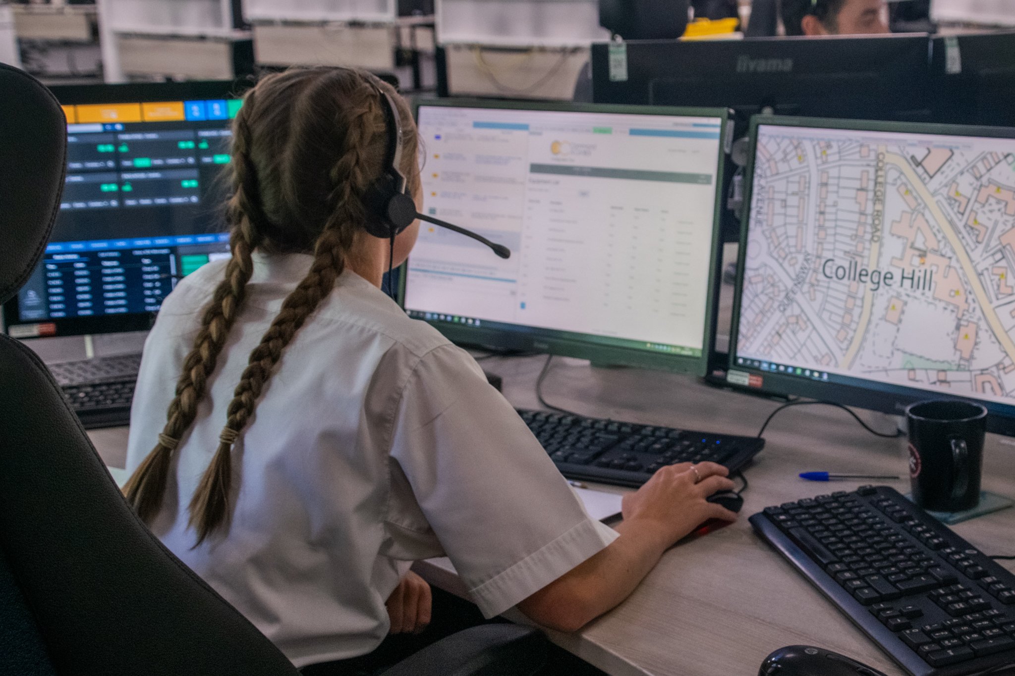 A woman with plaited hair wearing a headset works at a desk with multiple computer monitors showing maps and data in a control room.