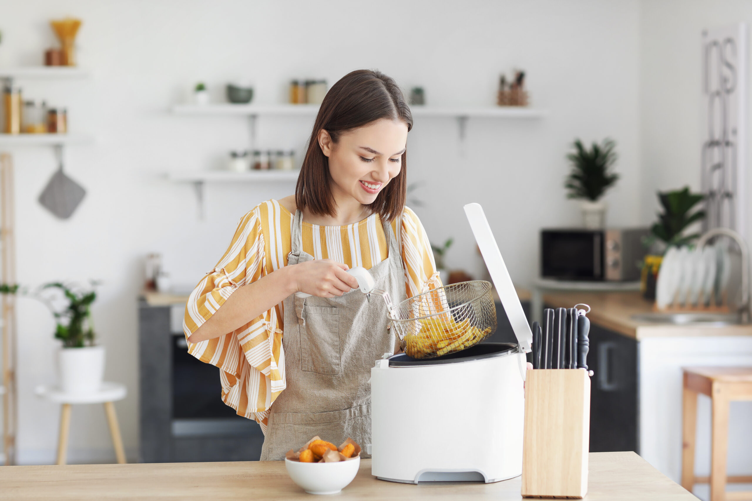 Young woman taking tasty french fries from deep fryer in kitchen
