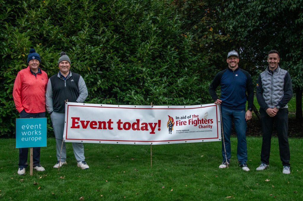 Four men in outdoor gear stand behind a banner for The Fire Fighters Charity reading ‘Event today!’. A sign for Event Works Europe is on the left.