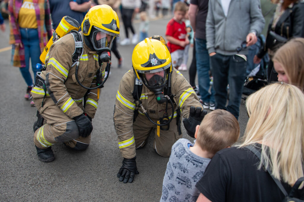 2 firefighters wearing full BA and PPE are kneeling down and greeting a young boy.