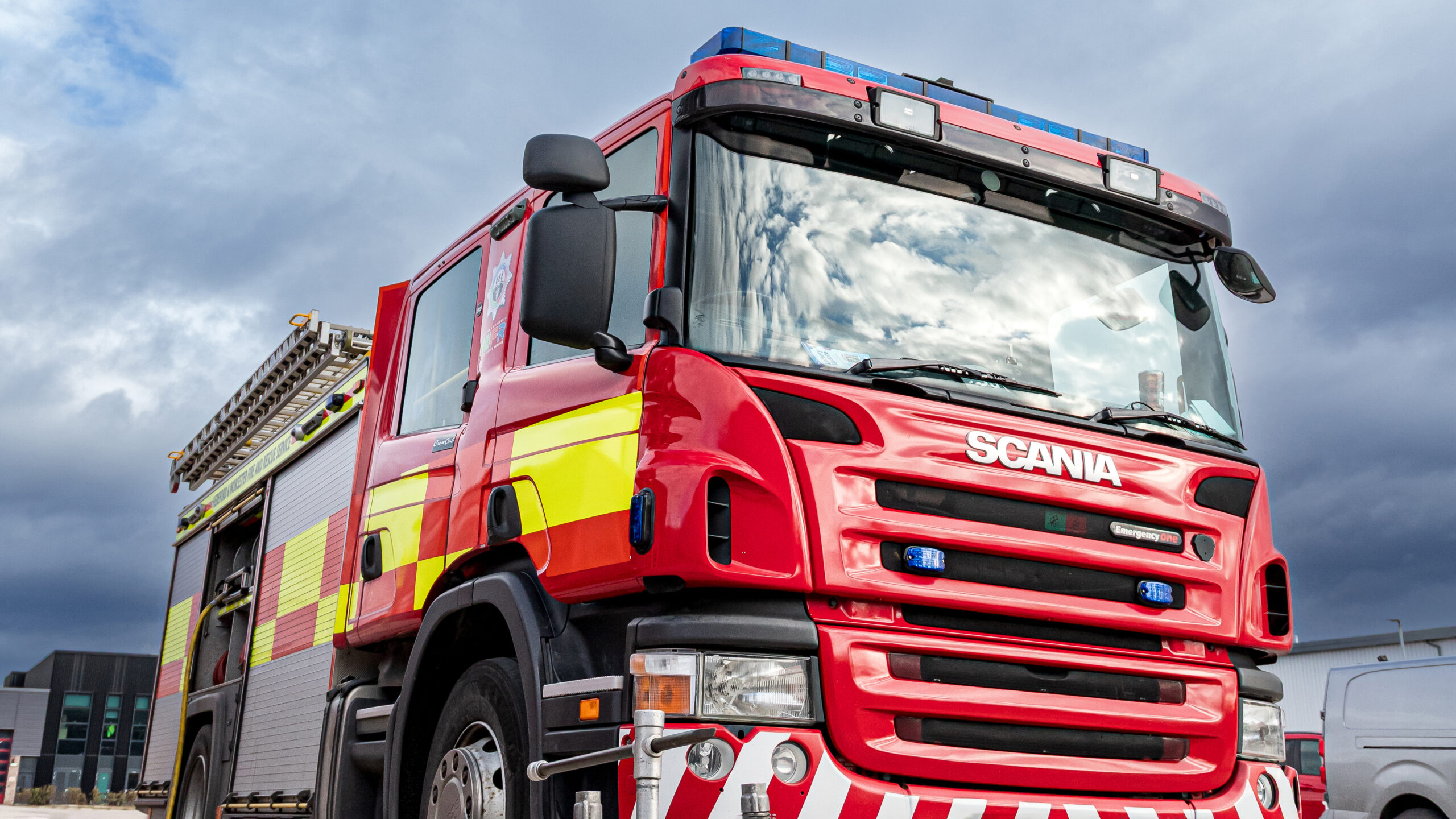 A red Scania fire engine from a low angle, featuring yellow high-visibility side markings and blue emergency lights under a cloudy sky.