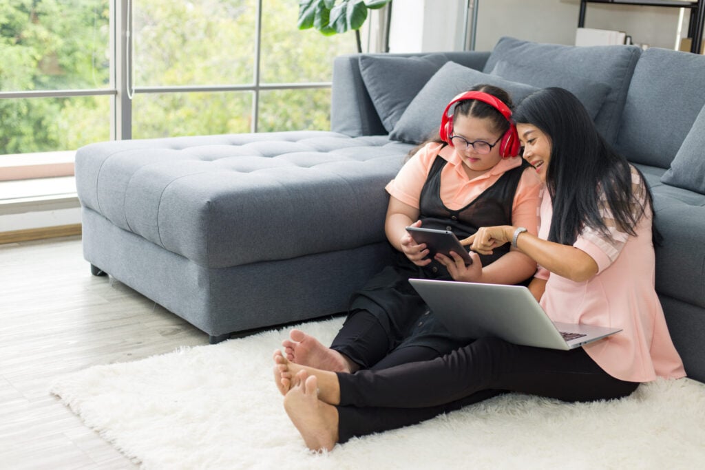 mother sitting on the floor next to her autistic daughter looking at an Ipad with headphones.