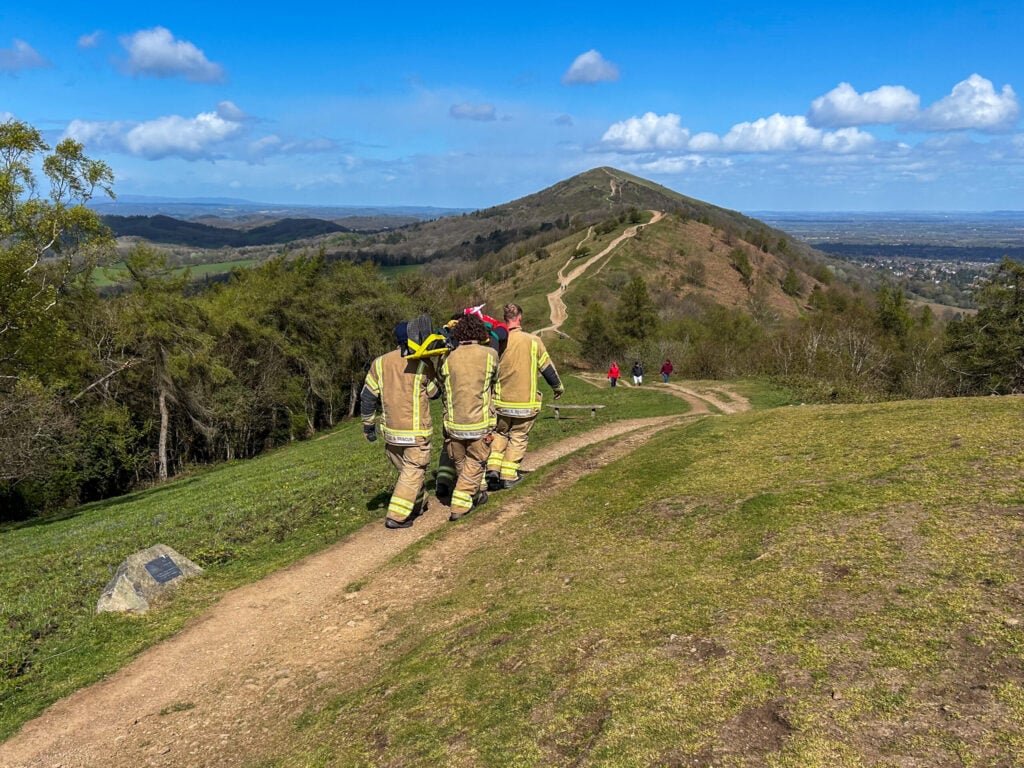 Four Firefighters carrying a casualty used in training across the Malvern Hills on a sunny day.